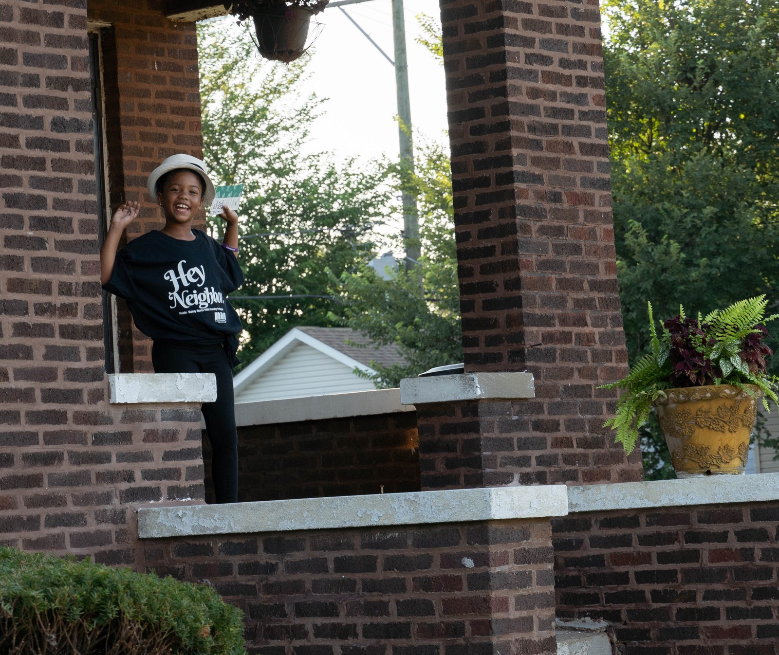 A smiling girl wearing a hat and Hey Neighbor shirt waves from a porch